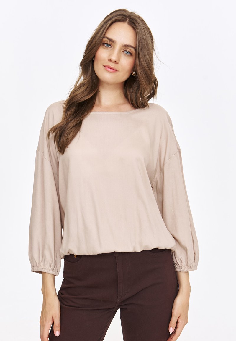 Beżowa, lekka bluzka o luźnym kroju, z dekoltem typu łódka i bufkami. Połączona z ciemnobrązowymi spodniami. Miękka tekstura i elegancki układ.