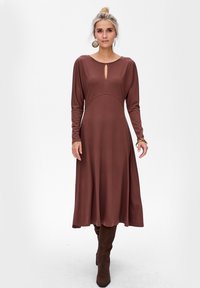 Robe marron à manches longues avec un col rond et un détail en trou de serrure, présentant un corsage ajusté et une jupe évasée. Porte des bottes marron au genou.