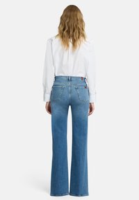 Niebieskie dzwoniaste jeansy z wysokim stanem, z pięcioma kieszeniami i subtelnym blaknięciem. Połączone z białą koszulą zapinaną na guziki.