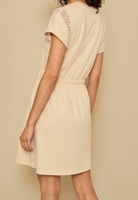 Robe beige courte au fit décontracté, taille élastique et accents en dentelle sur les épaules. Matière en coton doux avec une texture lisse.