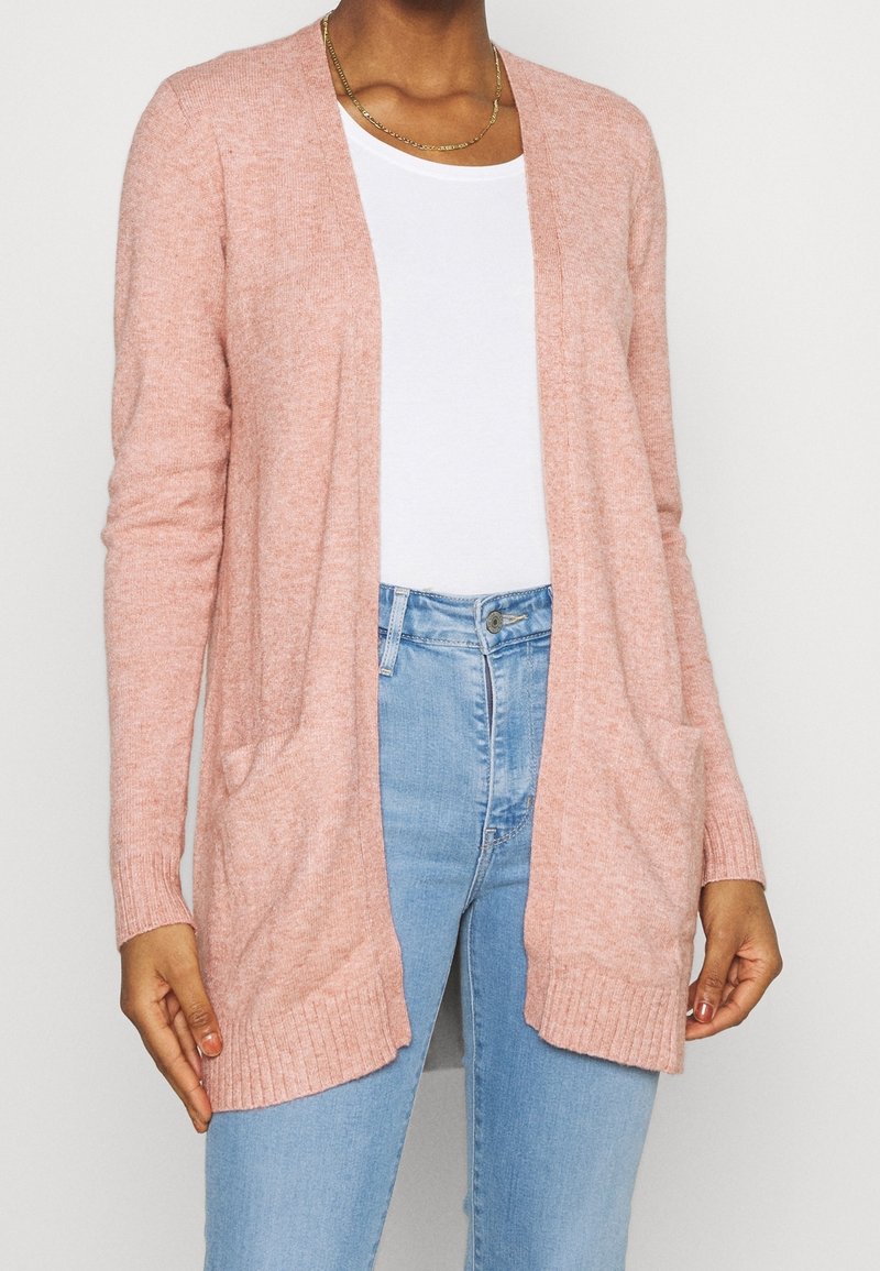 Femme portant un cardigan rose clair ouvert sur le devant par-dessus une chemise blanche et un jean bleu clair, debout devant un fond uni.