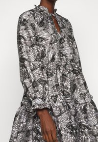 Grå mönstrad klänning med volanghalsringning och manschetter. Har abstrakta svarta och vita svängar, lagerdesign och lätt tyg.