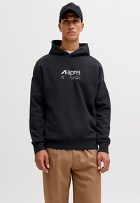 Moški z črno kapo, črno jopa s kapuco z belim napisom "aprel" in koordinatami ter rjavimi hlačami, stoji ob enobarvni sivi podlagi.