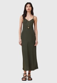 Vestido maxi verde oliva con tirantes tipo espagueti, de silueta ajustada y textura suave. Combinado con sandalias negras de tiras y un colgante largo.
