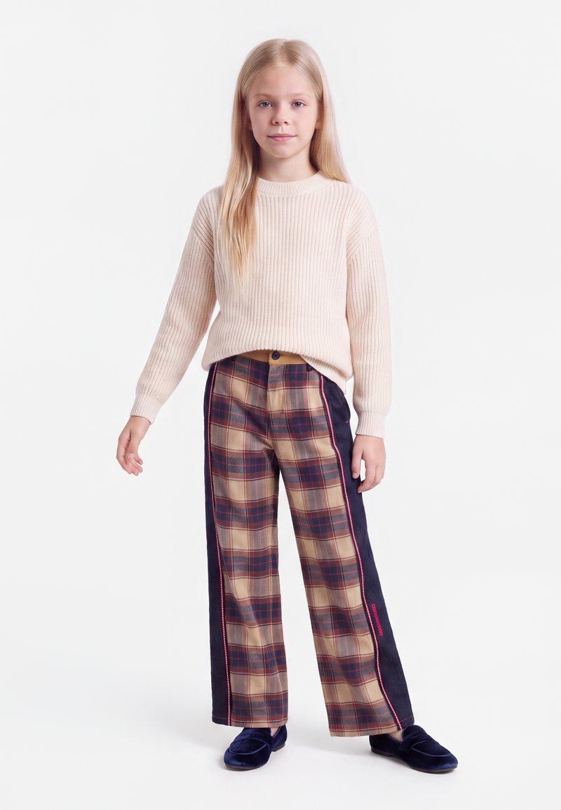 Fille blonde debout portant un pull côtelé beige clair, un pantalon large à carreaux avec des panneaux latéraux foncés et des chaussures en velours bleu foncé sur fond blanc.