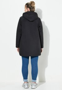 Schwarzer Kapuzenmantel mit geradem Schnitt, gestepptem Material und langen Ärmeln, kombiniert mit blauen Jeans und silbernen Sneakers.