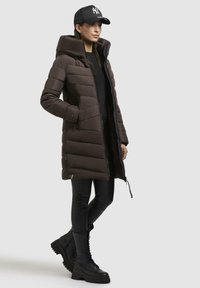 Giacca a vento marrone con colletto alto, chiusura con zip e design imbottito. Indossata sopra leggings neri e stivali neri con lacci chunky.
