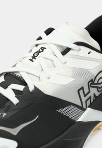 Αθλητικό παπούτσι ασπρόμαυρο με υφασμάτινο πάνω μέρος με λεπτομέρειες από δίχτυ, επενδεδυμένο κολάρο και κορδόνια σε αντίθεση. Το λογότυπο εμφανίζεται έντονα.