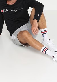 Schwarzes Langarmshirt mit "Champion"-Logo, graue Shorts, weiße Sneakers und gestreifte Socken mit roten und blauen Akzenten. Sitzt auf einer weißen Oberfläche.