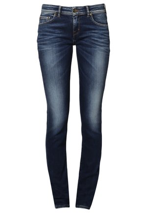 Femme portant un t-shirt blanc avec le texte « "Hilfiger Free Spirit" », un jean bleu, des baskets bleu marine et tenant des lunettes de soleil dans une main.