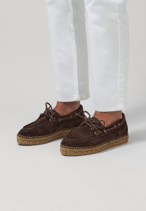 Brune ruskindssnørestøvler, espadrille, båret med hvide korte bukser på en lysegrå gulv.