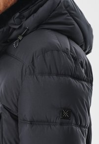 Czarna kurtka puchowa z kapturem, pikowanym wzorem i małą metką marki na rękawie. Szczegóły ze sznurkiem przy szyi.