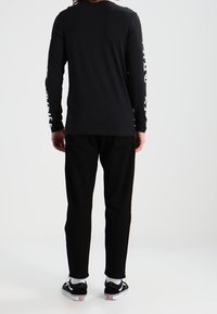 Black long-sleeve shirt with white printed text on sleeves, paired with black pants and black sneakers, viewed from the back.
