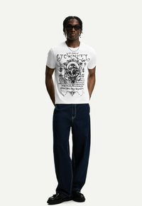T-shirt grafica bianca con testo e design stampati in nero sulla parte anteriore e posteriore, abbinata a jeans larghi in denim scuro e scarpe nere.