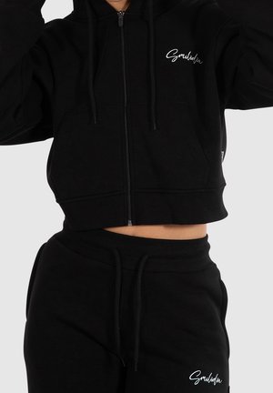 Person trägt schwarzen, kurz geschnittenen Kapuzenpullover mit Reißverschluss und passende schwarze Jogginghose, beide mit weißem "Smilodon" Logo-Schriftzug.