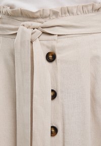 Beige pellavahame, jossa on sidottu vyötärö, röyhelövyötärö ja tummanruskeita nappeja etupaneelissa, esittelee teksturoitua kuitua.