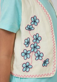 Bestickte Weste in Creme mit blauen Blumenmustern, umrandet in Pink. Getragen über einem türkisfarbenen Hemd aus einem weichen, strukturierten Stoff.