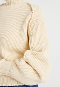 Pull en maille crème avec un col montant, présentant une texture côtelée, des manches froncées et un bord festonné le long de la couture de l'épaule.