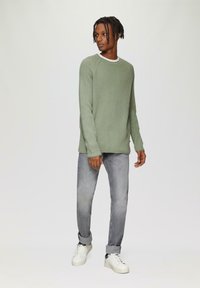 Grüner strukturierter Strickpullover mit einem weißen Kragen, kombiniert mit grauen, schmal zulaufenden Jeans und weißen Sneakern. Die Passform ist lässig und modern.