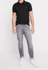 Homme portant un polo noir à manches courtes, un jean gris clair retroussé et des baskets montantes blanches, se tenant contre un fond blanc.