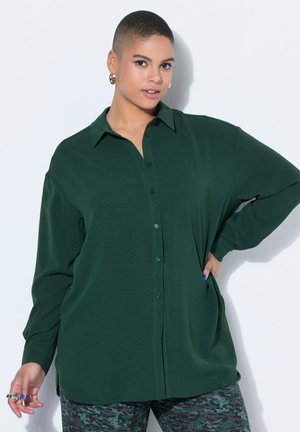 Sebevědomá osoba s krátkými vlasy, oblečená v tmavě zelené košili na knoflíky a v maskáčových kalhotách, pózující s rukou v bok proti bílému pozadí.