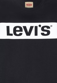 T-shirt nera in cotone con una evidente fascia orizzontale bianca recante il logo "Levi's" stampato in lettere nere in grassetto. Design classico con collo a giro.