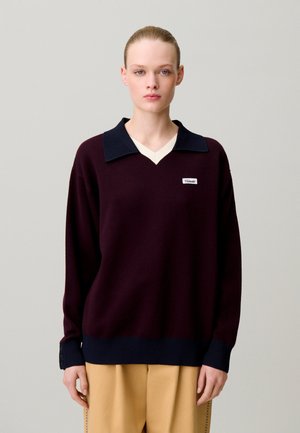 Jeune femme aux cheveux tirés en arrière portant un pull bordeaux foncé avec un col noir et un pantalon beige sur un fond uni.