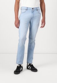 Lichtblauwe spijkerbroek met een rechte snit, vijf zakken en een metalen knoopsluiting. Draagt een zwart paar sneakers en een wit shirt.