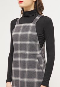 Robe salopette à carreaux gris en tissu texturé, avec de larges bretelles, une poche latérale, portée avec un haut noir à manches longues.