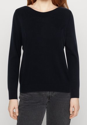 Vrouw die een effen zwarte trui met lange mouwen en een ronde hals draagt en donkere grijze jeans, afgebeeld van de nek tot aan de heupen.
