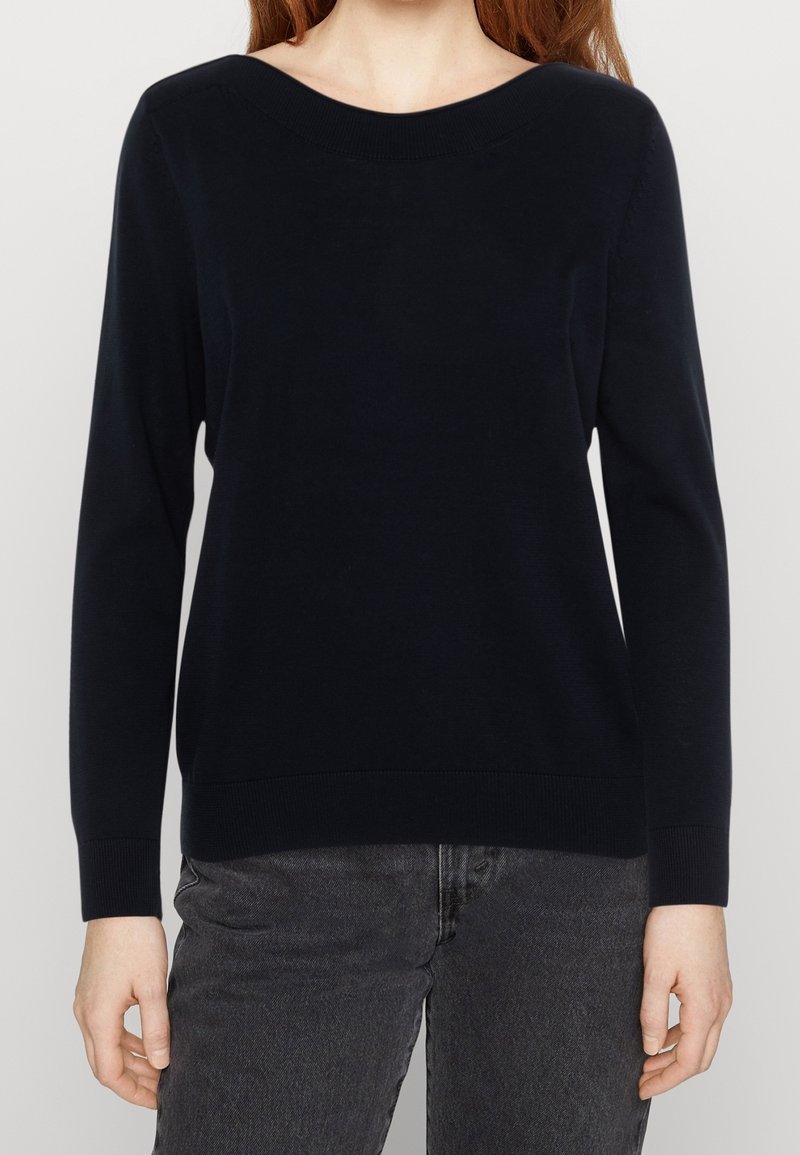 Femme portant un pull noir uni à manches longues avec un col rond et un jean gris foncé, montrée du cou jusqu’aux hanches.