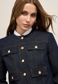 Giacca in denim scuro con un design strutturato, dotata di bottoni a pressione in tonalità dorata, doppie tasche sul petto e cuciture a contrasto.