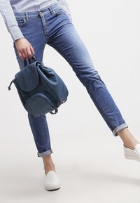 Niebieski skórzany plecak z zaokrąglonymi kieszeniami, miękką fakturą i zakończeniem na sznurek. Założony z jasnoniebieskimi spodniami typu jeansy z podwiniętymi nogawkami i białymi butami typu slip-on.