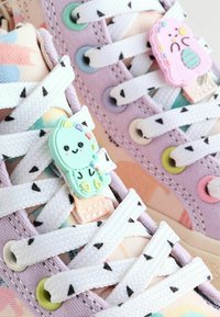 Le sneakers in tela multicolore presentano lacci bianchi con due clip a forma di personaggi, accenti pastello e una superficie in tessuto testurizzato con motivi vivaci.