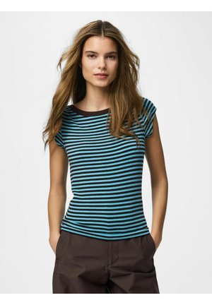 Kvinde med langt hår iført en blå- og sortstribet kortærmet skjorte og mørke bukser, står med hænderne i lommerne mod hvid baggrund.