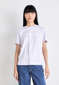 Fialové bavlněné tričko s krátkým rukávem, na kterém se nachází bílé tištěné logo "Calvin Klein Jeans" na prsou. Volný střih.
