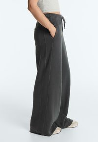 Person trägt dunkelgraue, weit geschnittene Jogginghosen mit Taschen vorne und hinten, kombiniert mit einem hellgrauen, cropped Oberteil und beigen Turnschuhen.