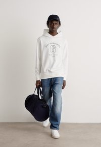 Sudadera blanca con logo, jeans azules, zapatillas blancas y bolsa de duffel azul marino. El modelo lleva una gorra azul marino, de pie contra un fondo neutral.