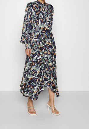 Femme portant une longue robe fluide avec des motifs de feuilles vertes et orange, boutonnée sur le devant, manches bouffantes et sandales blanches à talons hauts.