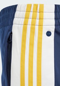 Námorníkové sportovní kraťasy s bílými a žlutými pruhy, obsahující zipovou kapsu a strukturovaný pás. Zahrnuje malou modrou logo akcentaci.