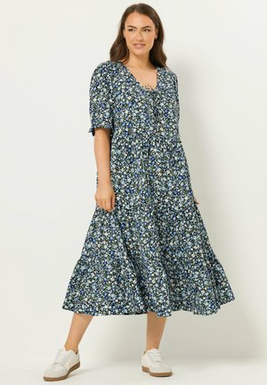 Kvinde i en blå midikjole med blomstermønster, lagdelt skørt, korte ærmer og hvide sneakers, stående foran en ensartet lys baggrund.