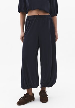 Femme portant un pantalon ample bleu marine court avec une ceinture élastique et des baskets marron à lacets, debout devant un fond blanc.