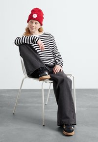 Chemise à rayures noir et blanc à manches longues, jeans noirs à jambe large, bonnet en tricot rouge avec logo, et baskets noires avec semelles en caoutchouc sur une chaise blanche.