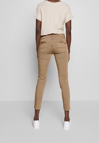 Person som bär beige slim-fit byxor med bakfickor, en ljusbeige t-shirt med kort ärm och vita sneakers står mot en grå bakgrund.