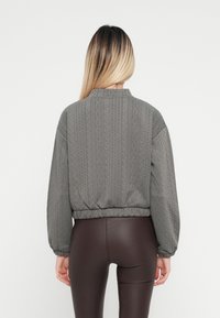Veste en tricot grise avec un motif texturé, ourlet élastique et manches ballons, associée à des leggings marron. Vue de dos ; design minimaliste.