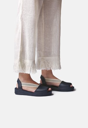 Personne portant des sandales compensées noires à bout ouvert avec des bandes élastiques beige rayées et un pantalon blanc à jambes larges avec ourlets à franges.