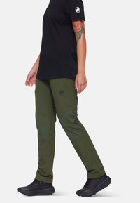 Olijfgroene outdoorbroek met een taps toelopende pasvorm, voorzien van donkerdere groene paneelaccenten en een logo. Draagt een zwart shirt met korte mouwen en zwarte schoenen.