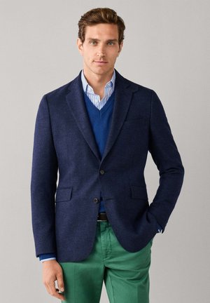 Man draagt een navy blazer, een blauwe V-hals trui, een gestreept blauw overhemd en groene broek, staand met één hand in de zak tegen een grijze achtergrond.
