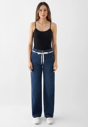 Woman with long hair stands facing forward wearing black tank top, wide-leg blue jeans with white tie belt, and white sneakers against white background.