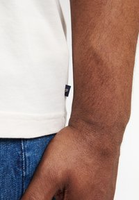 Camiseta blanca de algodón de manga corta con una pequeña etiqueta azul en el dobladillo, combinada con jeans de mezclilla azul, mostrando una mano descansando sobre el muslo.